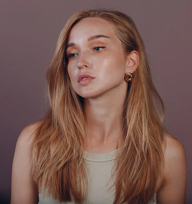 A young woman with shoulder-length light brown hair poses against a dusty rose background. She has soft makeup with a natural pink lip and light-toned skin. Wearing a beige tank top, she accessorizes with a gold circular earring. The lighting is soft, highlighting her face and upper body. - Cat Eye Reshaping in Houston, TX
