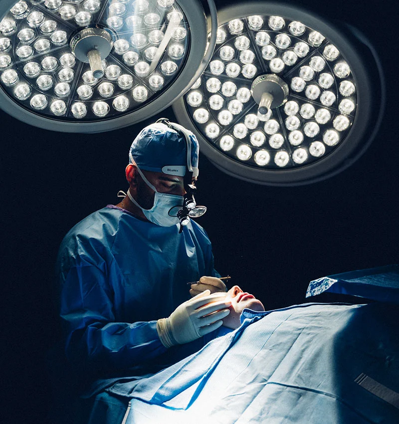 A full indoor shot of Dr. Chattha performing surgery, dressed in a light-blue gown, cap, and mask. He wears white surgical gloves and holds a small instrument near the patient’s face, who is draped in a matching surgical cover. Two large, bright circular lights overhead illuminate the operating area, contrasting with the darker surroundings. The scene emphasizes the sterile, controlled environment and the precision of the procedure. - Revision Facelift in Houston, TX